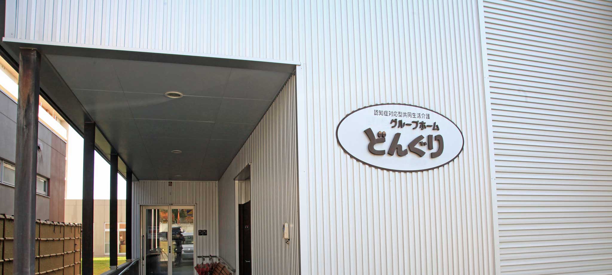 施設の玄関・車寄せ。白い金属製外壁に施設名「どんぐり」の看板が見える